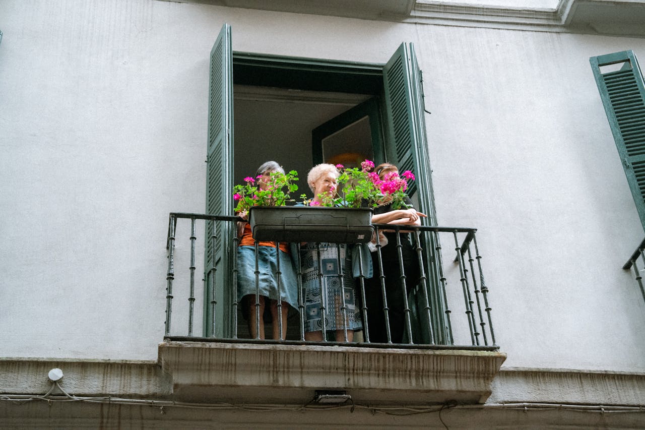 An elderly woman standing on a balcony adorned with vibrant flowers, enjoying the view outdoors.
